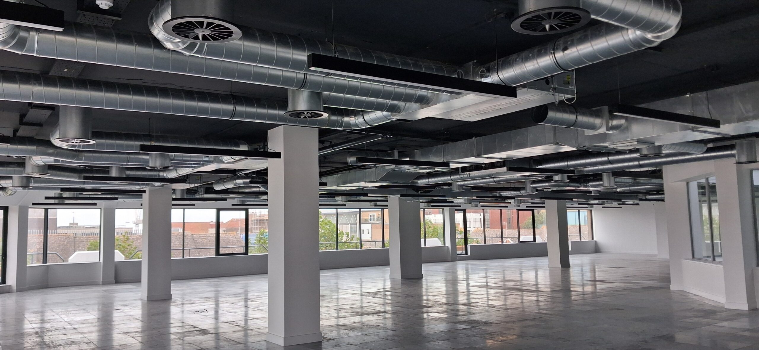 Empty modern office space with large windows, tiled floors, white walls, and exposed metal ductwork on the black ceiling.