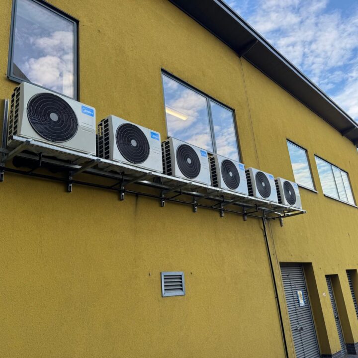 Six Midea air conditioning units are mounted in a row on the exterior wall of a yellow building with large windows, set against a clear blue sky in the background.