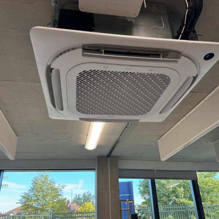 A ceiling-mounted Midea air conditioning unit with vents is installed in an office with large windows and views of trees and people outside.