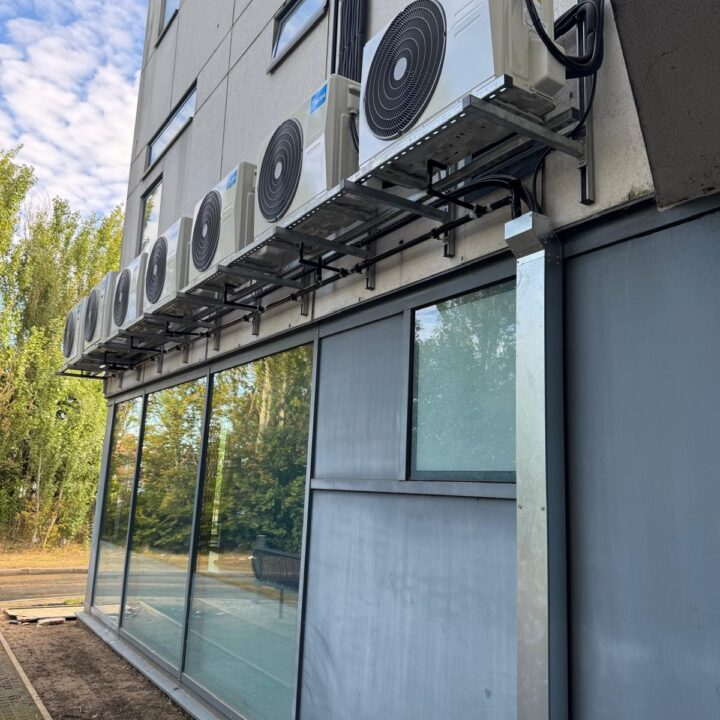 Six Midea air conditioning units are mounted on a metal rack attached to the exterior wall of a modern building with large windows.