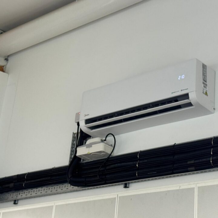 A Midea air conditioning unit is wall-mounted above exposed black cables on a white wall, displaying a temperature of 20 degrees Celsius.