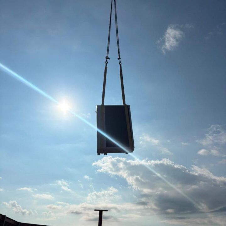 A large rectangular object is suspended by cables from a crane against a bright sky with scattered clouds and the sun shining.